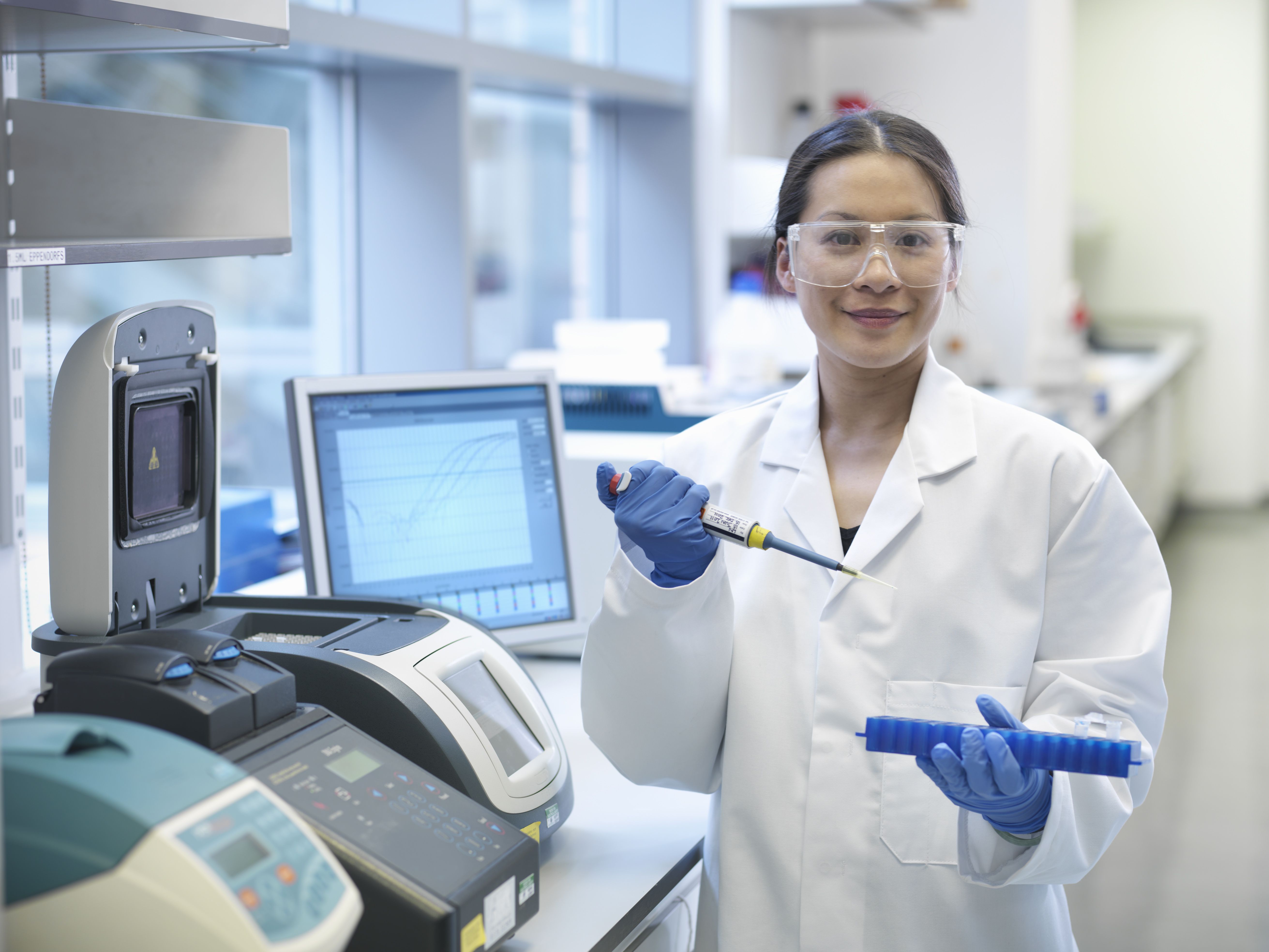Woman in a lab coat and safety goggles holding a micropipetter.